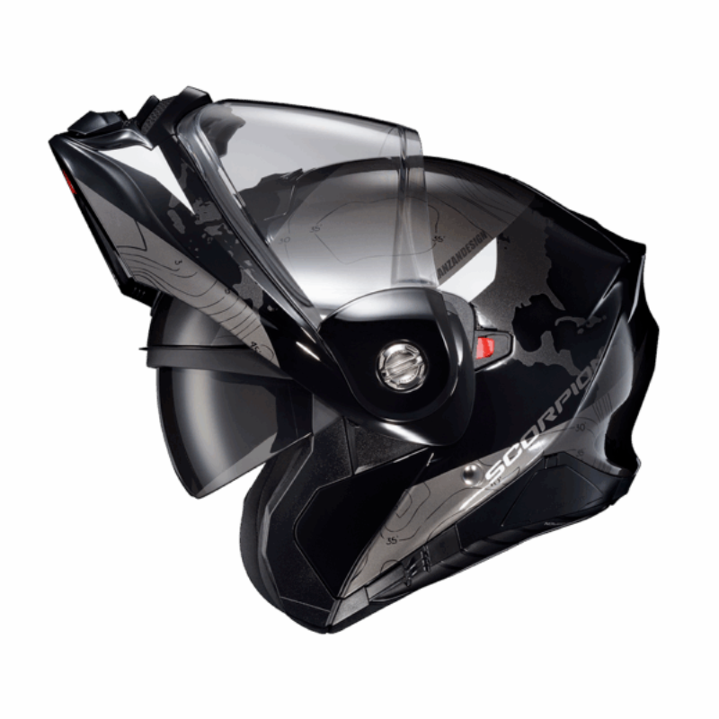 Side profile of the Scorpion EXO-AT960 Topographic Modular Adventure Motorcycle Helmet in Black/White with the modular chin bar in the fully raised position. The helmet features a matte black and metallic grey shell with topographic map graphics and white accent panels. Visible are the clear EverClear® face shield, integrated SpeedView® drop-down sun visor, and the convenient flip-up chin bar that allows easy communication, fueling, and improved comfort when stopped. Designed for adventure touring and dual-