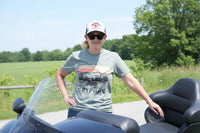 Person wearing a cap and sunglasses wearing a green Gryphon Any Road Any Ride T-shirt outdoors with greenery in the background