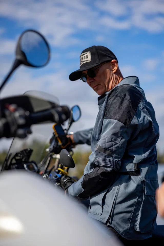 Man in a black cap and gray jacket standing next to a motorcycle with a blue sky background