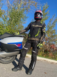 Person in motorcycle gear standing next to a motorcycle on a road with trees in the background
