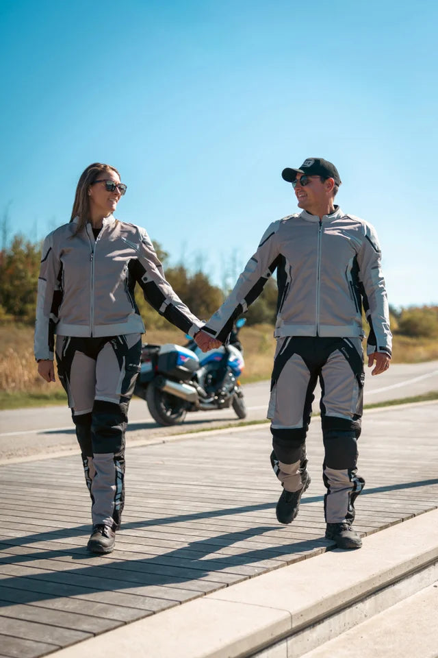 Two people in motorcycle gear walking on a road with a motorcycle in the background.