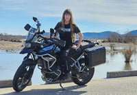 Woman standing on a motorcycle wearing a black Gryphon branded T-shirt by a water body with mountains in the background