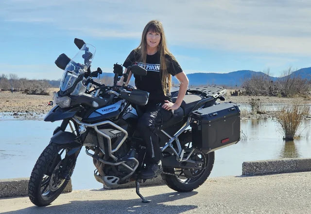 Woman standing on a motorcycle wearing a black Gryphon branded T-shirt by a water body with mountains in the background