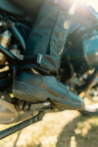 Close-up of Gryphon Women’s Pacific Coast 2 black motorcycle pants with an adjustable ankle closure worn over a riding boot on a motorcycle footpeg.