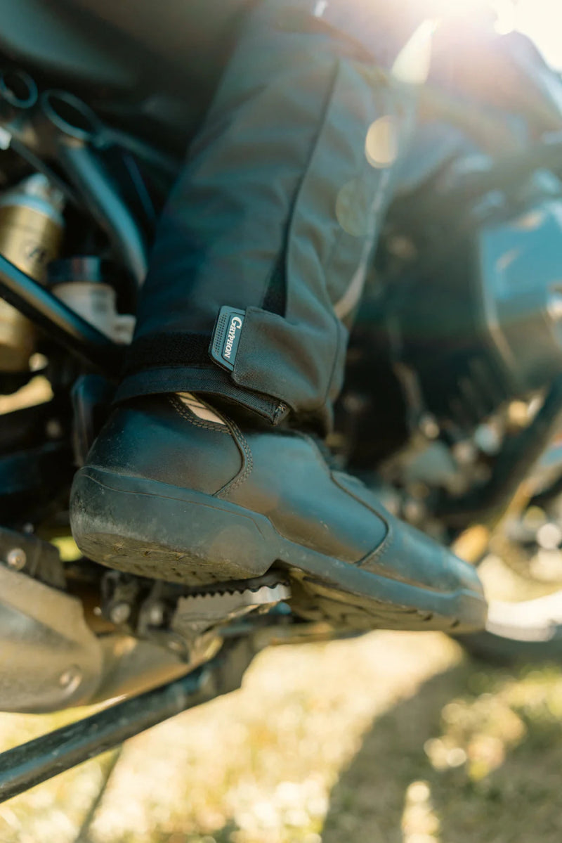 Close-up of Gryphon Women’s Pacific Coast 2 black motorcycle pants with an adjustable ankle closure worn over a riding boot on a motorcycle footpeg.