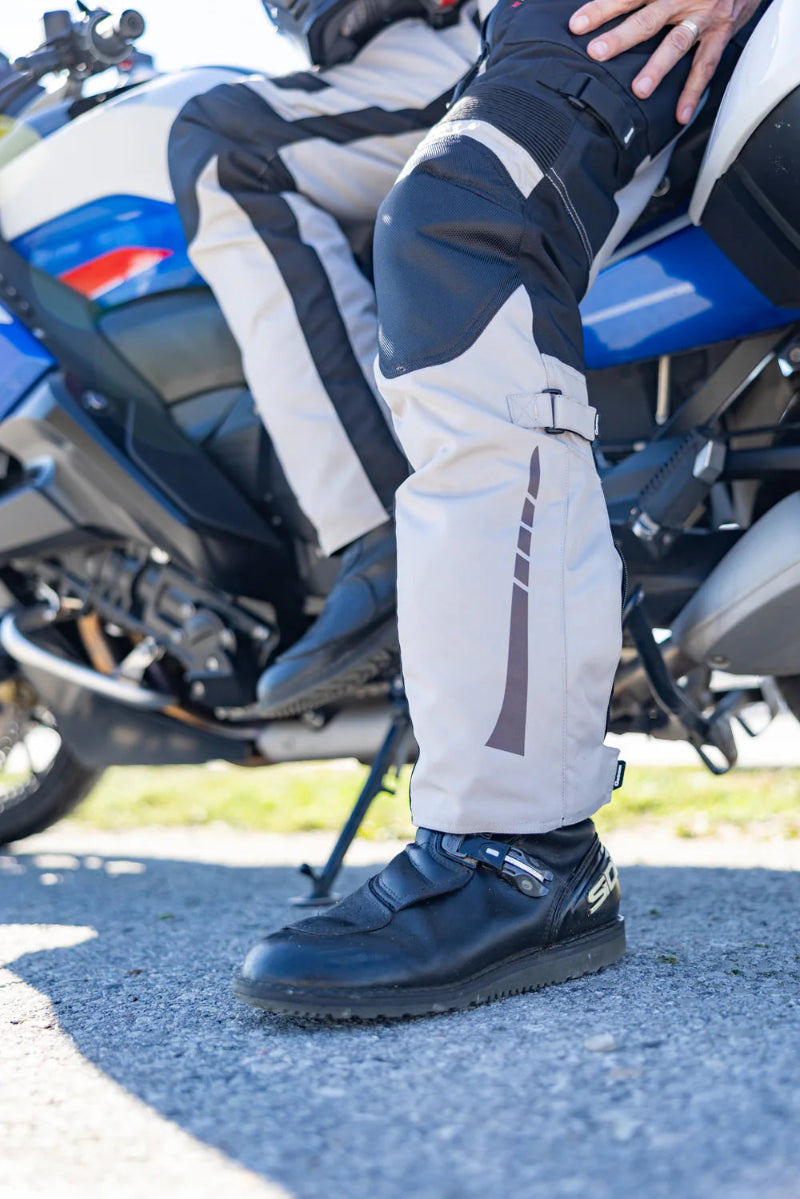 Woman wearing Gryphon Pacific Coast 2 grey and black adventure motorcycle pants with reinforced knees and reflective lower-leg detailing beside a motorcycle.