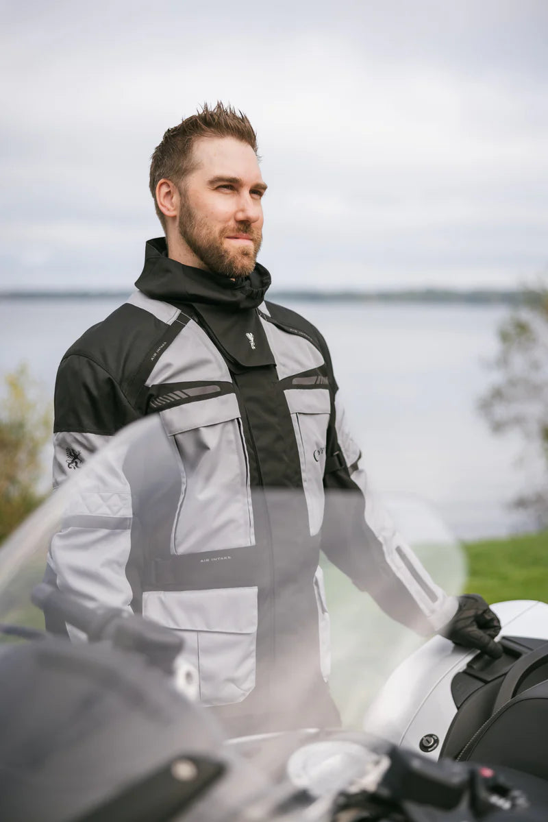 Man wearing a black and gray jacket standing next to a motorcycle by a lake.