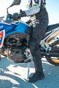 Woman wearing Gryphon Women’s Pacific Coast 2 black waterproof motorcycle pants with adjustable leg straps and reflective detailing while seated on an adventure motorcycle.