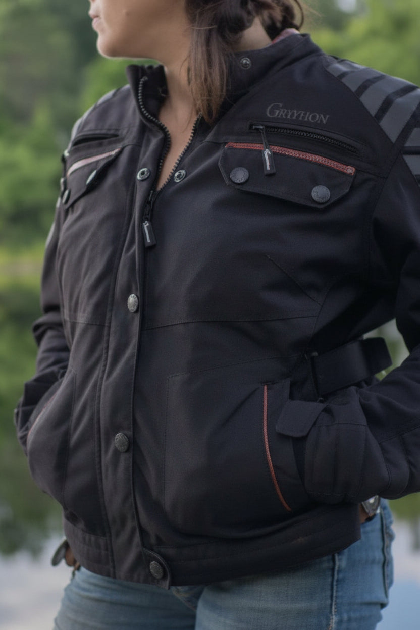 Woman wearing a Gryphon Women’s Rebel black waterproof motorcycle jacket with reflective shoulder panels, red accents and adjustable waist straps.