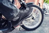 Lifestyle photo of the Joe Rocket Blaster Waterproof Motorcycle Shoes being worn while riding a motorcycle. The black suede motorcycle shoes feature a reinforced rubber shift pad, lace-up design, protective ankle support, and sneaker-inspired styling, providing waterproof comfort, grip, and protection for commuting, urban riding, and everyday motorcycle adventures.