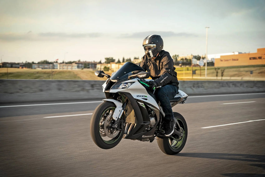 A rider wearing a black helmet and leather riding gear maneuvers a white and black sport motorcycle on an open road during golden hour.