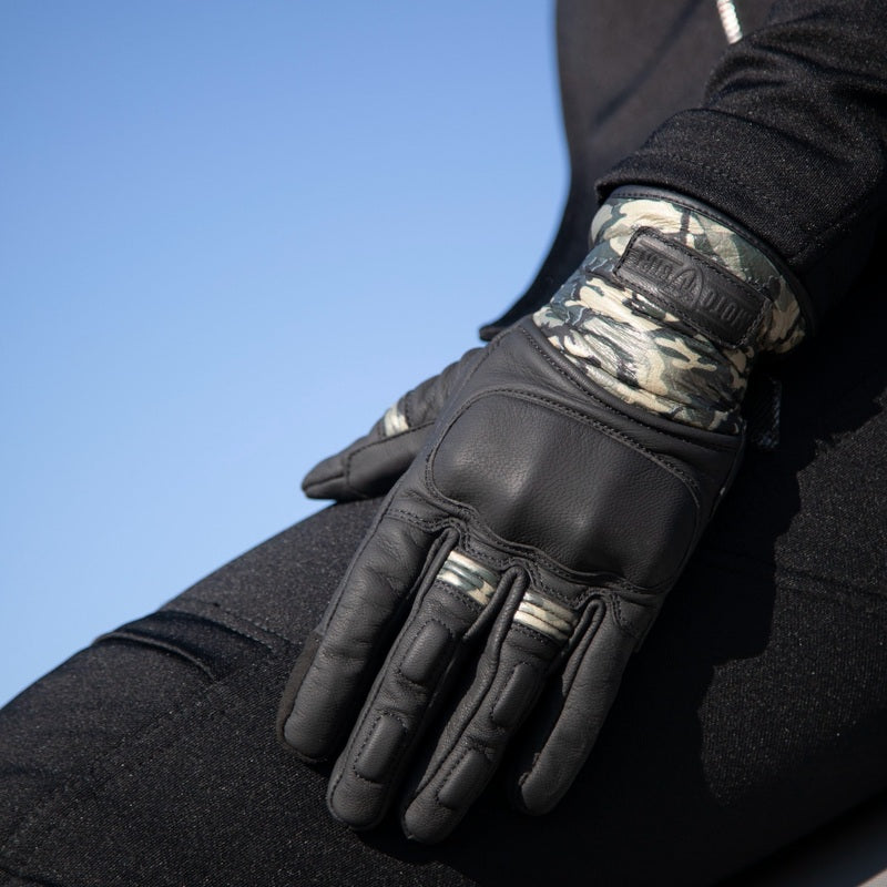 Close-up of MotoGirl Bessie black leather motorcycle glove worn by a rider, showing padded knuckle protection, reinforced fingers and camouflage wrist detailing.