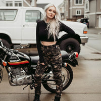 Person standing in front of a motorcycle wearing Motogirl branded Lara Cargo motorcycle pants in camo and a black cropped long sleeve shirt