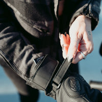 Woman fastening the adjustable hook-and-loop wrist closure on a black MotoGirl Nandi motorcycle glove, showing knuckle protection and secure fastening.