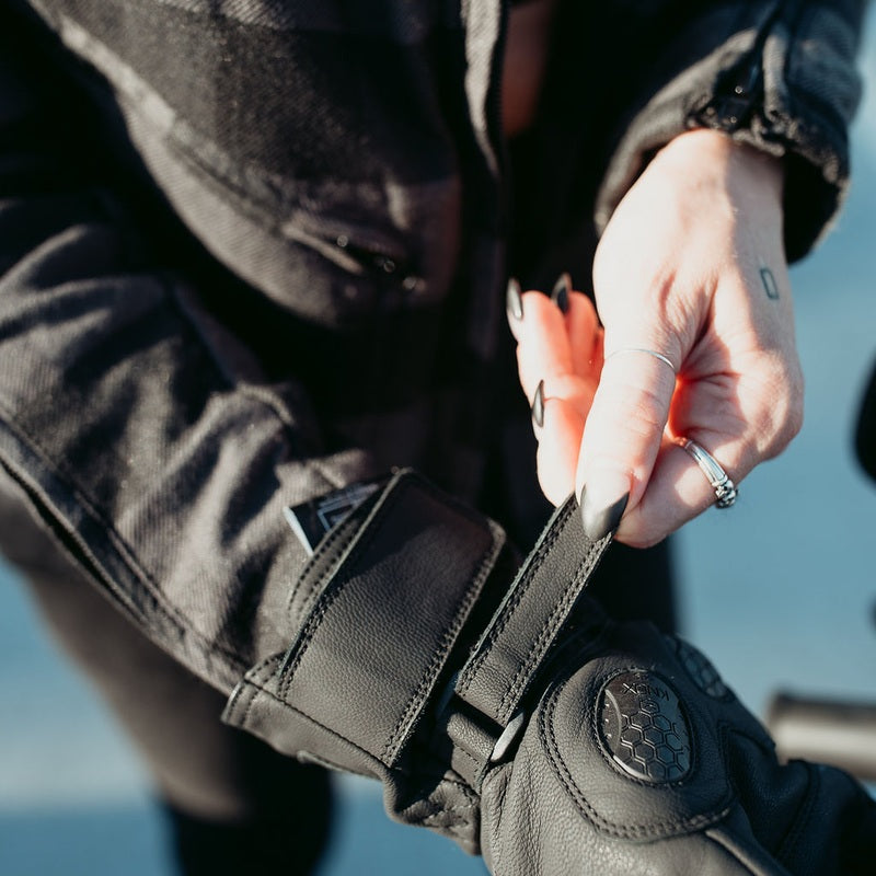 Woman fastening the adjustable hook-and-loop wrist closure on a black MotoGirl Nandi motorcycle glove, showing knuckle protection and secure fastening.