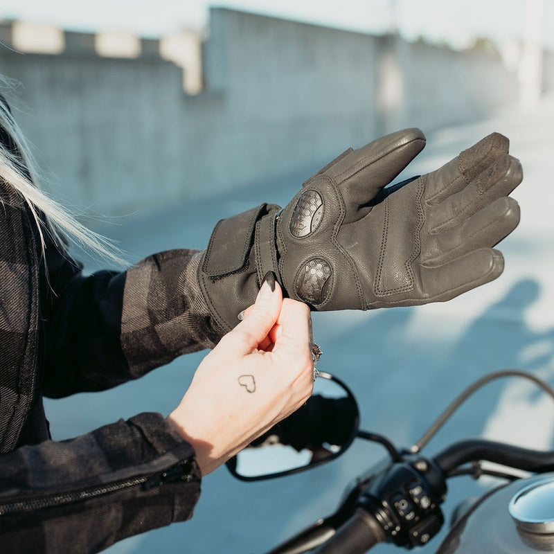 Woman adjusting black MotoGirl Nandi motorcycle glove beside a motorcycle, showing protective knuckle panels, palm construction and adjustable wrist fastening.
