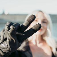 Close-up of black MotoGirl Nandi motorcycle glove showing touchscreen-compatible index and middle fingertips with a woman in the background.