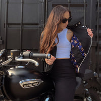 a girl wearing a white tank op and a Motogriel branded sherrie motorcycle jacket in black showing the inside colourful liner and zipper sitting on a motorcycle with a metal storage container in the background