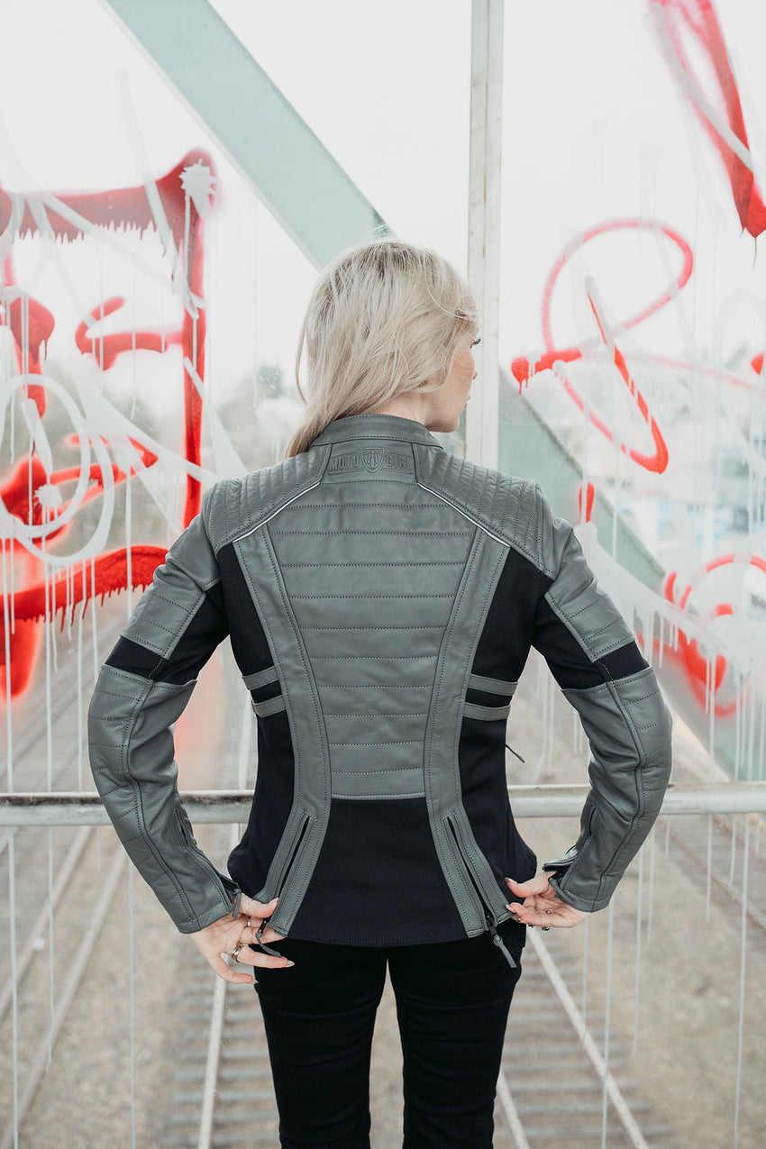 a girl earing a Motogirl Amelia Motorcycle Jacket in Grey with black accents and visible hip adjuster zippers with graffiti in the background