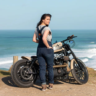 A girl wearing a grey t-shirt with blue denim overalls and brown sneakers in front of a motorcycle and the sea in the background