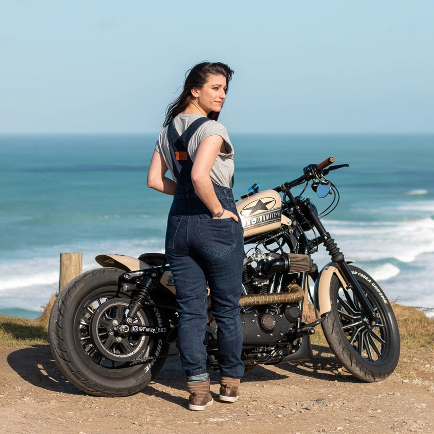 A girl wearing a grey t-shirt with blue denim overalls and brown sneakers in front of a motorcycle and the sea in the background