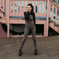 A person wearing a long sleeve black short with Motogirl branded grey denim overalls paired with black shoes with stairs in the background