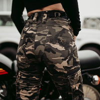 Camouflage pants with a visible brand logo worn by a person, with a motorcycle in the background.