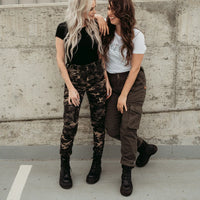 Two women standing together against a concrete wall, wearing casual outfits with camouflage pants and boots.