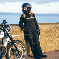 Lifestyle photo of a rider wearing the MotoGirl Vanessa Women's Motorcycle Jacket and Vanessa Motorcycle Pants in black while leaning against a seaside wall beside a motorcycle. The outfit features reflective orange accents, cargo pockets, and an adventure-ready design, combining waterproof performance, CE armour protection, and all-day comfort for touring, commuting, and long-distance motorcycle riding.