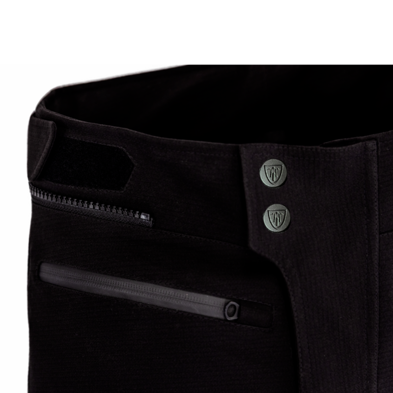 Close-up of the MotoGirl Vanessa Women's Motorcycle Pants waistband showing the secure dual-snap front closure, hook-and-loop waist adjustment, and waterproof zippered front pockets. The durable black textile construction provides a comfortable, adjustable fit for touring, adventure, and everyday motorcycle riding.