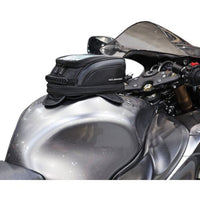 A black motorcycle tank bag attached to the fuel tank of a motorcycle, featuring a dual mounting system with visible straps and a waterproof rain cover.