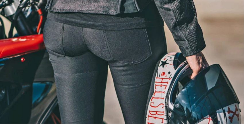 A person wearing dark gray biker shorts and a dark jacket stands beside a motorcycle, holding a helmet with a red, white, and black graphic design.