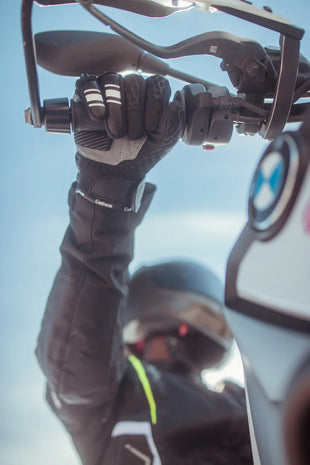 Rider wearing Gryphon Sahara Vented Motorcycle Gloves while gripping the handlebar of an adventure motorcycle. The black and grey summer riding glove features breathable 3D mesh construction, TPU knuckle protection, reinforced leather palm, and touchscreen-compatible fingertips. Shown in a real-world riding position, the glove provides excellent grip, ventilation, and comfort for warm-weather street and adventure motorcycle riding.