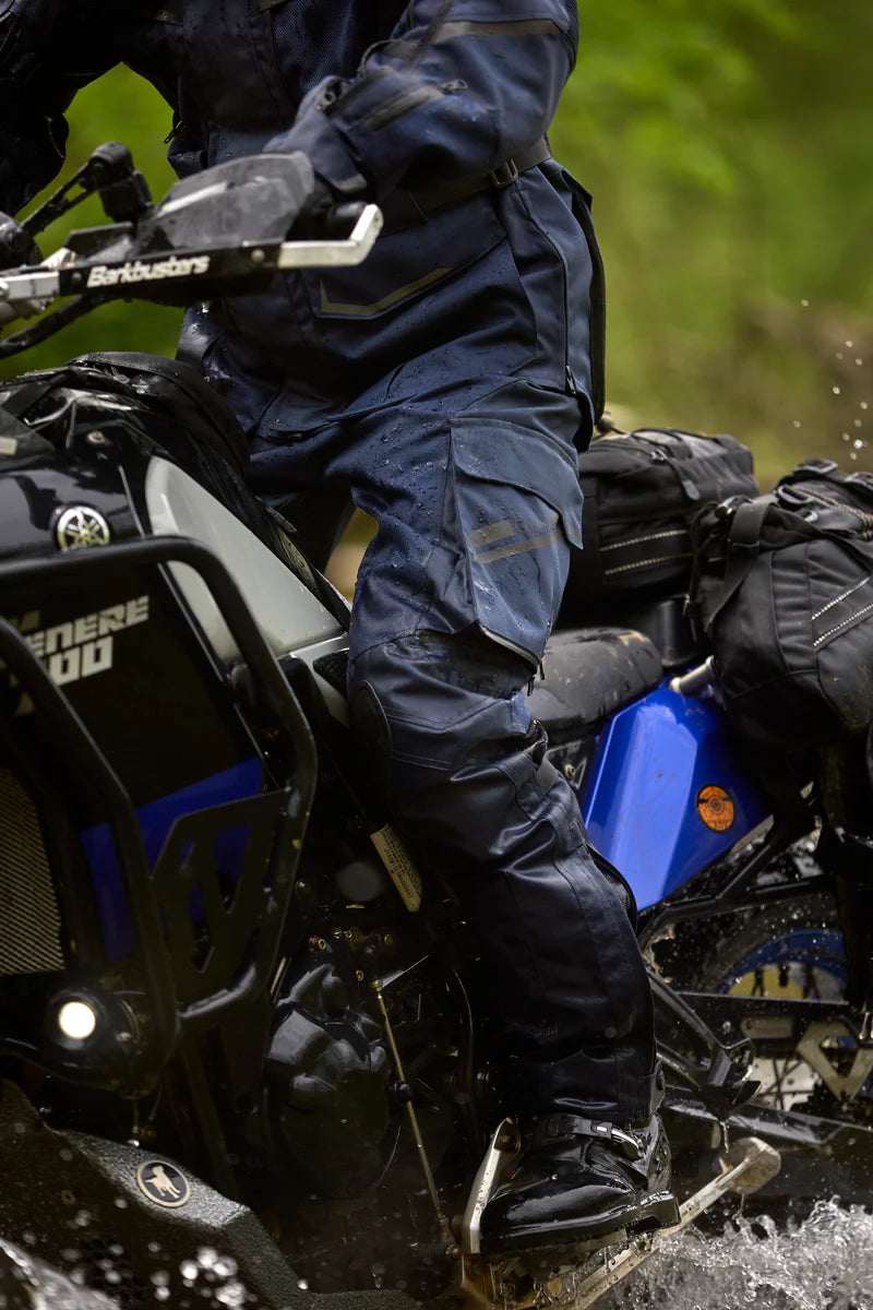 Rider wearing Gryphon Single Track Waterproof Motorcycle Pants and matching adventure jacket while riding a Yamaha Ténéré 700 through water on an off-road trail. The blue waterproof pants feature reinforced knee protection, cargo pockets, adjustable lower legs, and a durable adventure-ready design built for long-distance touring, dual-sport riding, and rugged off-road conditions.