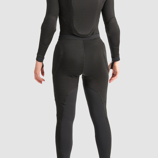 Back view of a woman wearing a black motorcycle protective base layer suit, showing the integrated back protector, elbow protection, fitted leggings, and ankle stirrups against a light grey background.