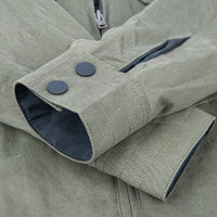 Close-up of an olive green jacket sleeve with two dark snap buttons, a dark blue inner lining, and a zipper, shown against a matching fabric background.