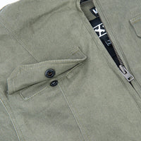 Close-up of an olive green canvas jacket with a black buttoned flap pocket, a silver zipper, and a black neck label with white text.