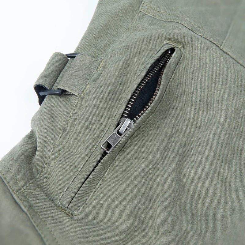 Close-up of an olive green canvas utility pocket with a diagonal zipper and silver metal pull, shown on a light gray background.