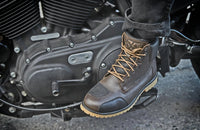 Lifestyle image of the Speed and Strength CALL TO ARMS Motorcycle Boots in brown and black being worn on a motorcycle. The premium leather riding boots feature a reinforced toe shift pad, padded ankle collar, lace-up design, and rugged gum outsole, combining classic work-boot styling with dependable protection and all-day comfort for commuting, cruising, and everyday riding.