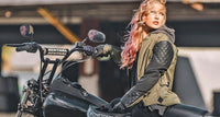 A woman with pink hair sits on a black motorcycle in a rustic indoor setting. She is wearing an olive green quilted vest over a black long-sleeve shirt and black leather gloves.