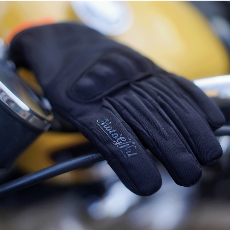 Close-up lifestyle photo of the MotoGirl Amina Women's Motorcycle Glove resting on a motorcycle handlebar. The glove features a soft textile upper, subtle knuckle protection, premium leather palm, touchscreen-compatible fingertips, and embroidered MotoGirl branding on the index finger. Designed for women riders seeking lightweight comfort, everyday protection, and classic motorcycle style.